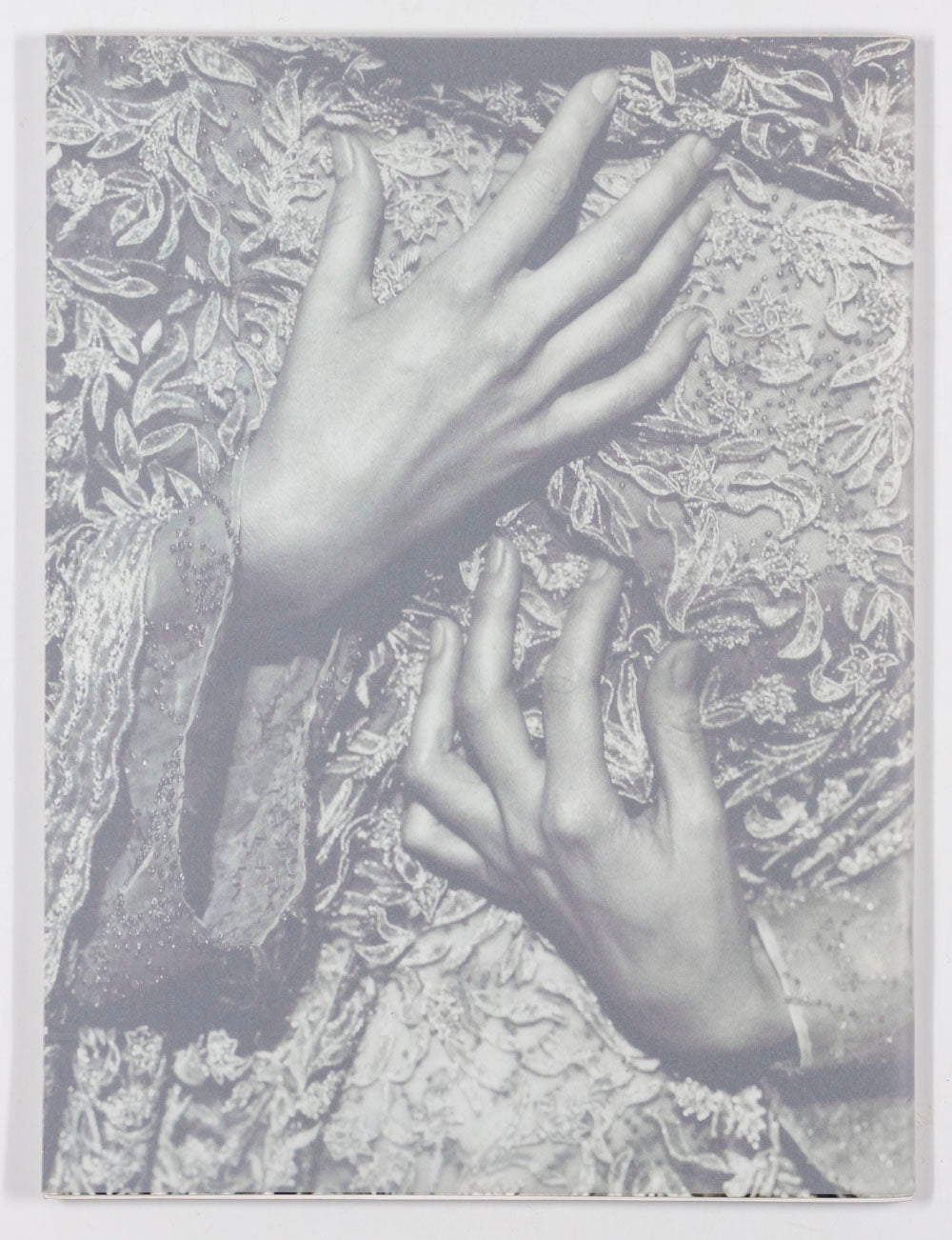 A monochrome artistic photograph featuring two hands resting on ornate, textured lace fabric.