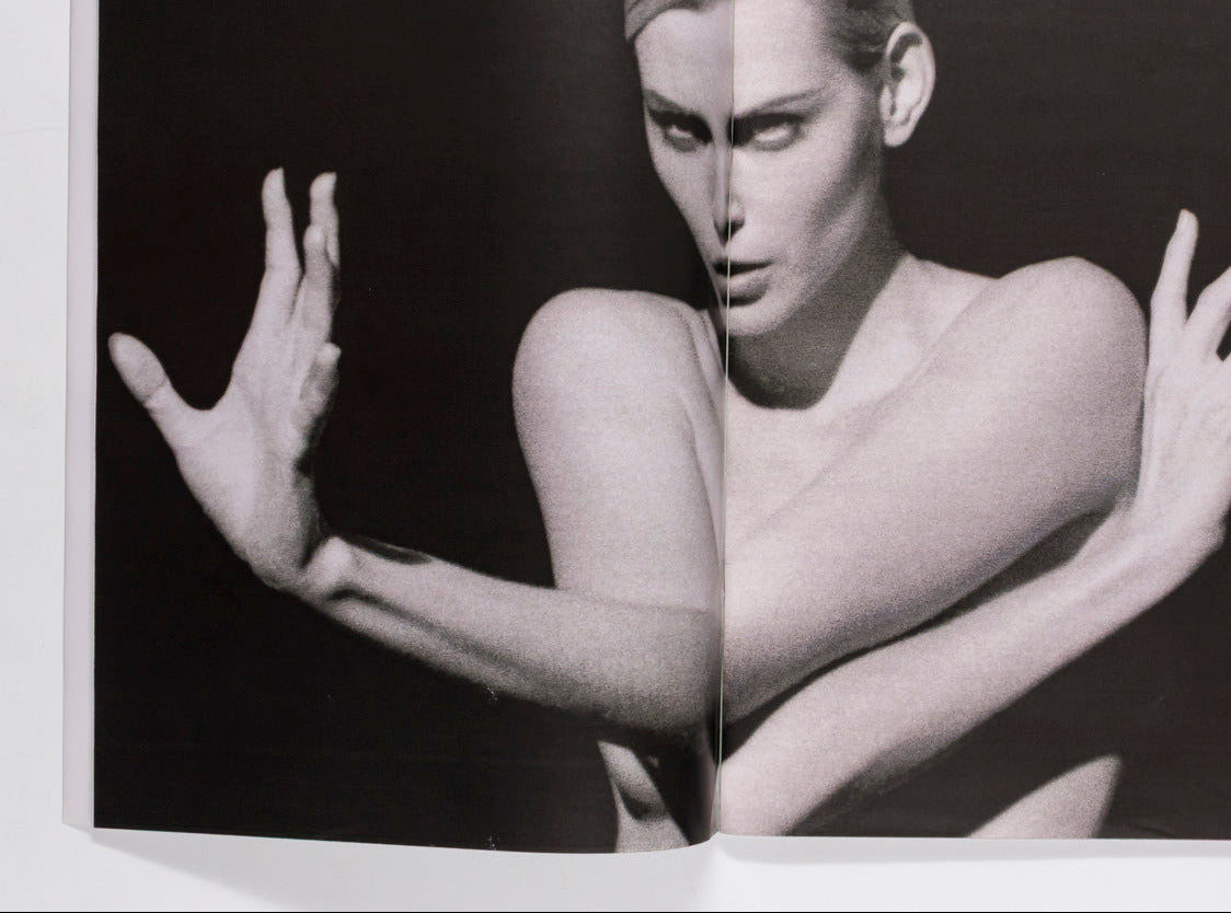 An open book or magazine spread showing a black-and-white fashion photograph of a nude woman with arms raised against a dark background.