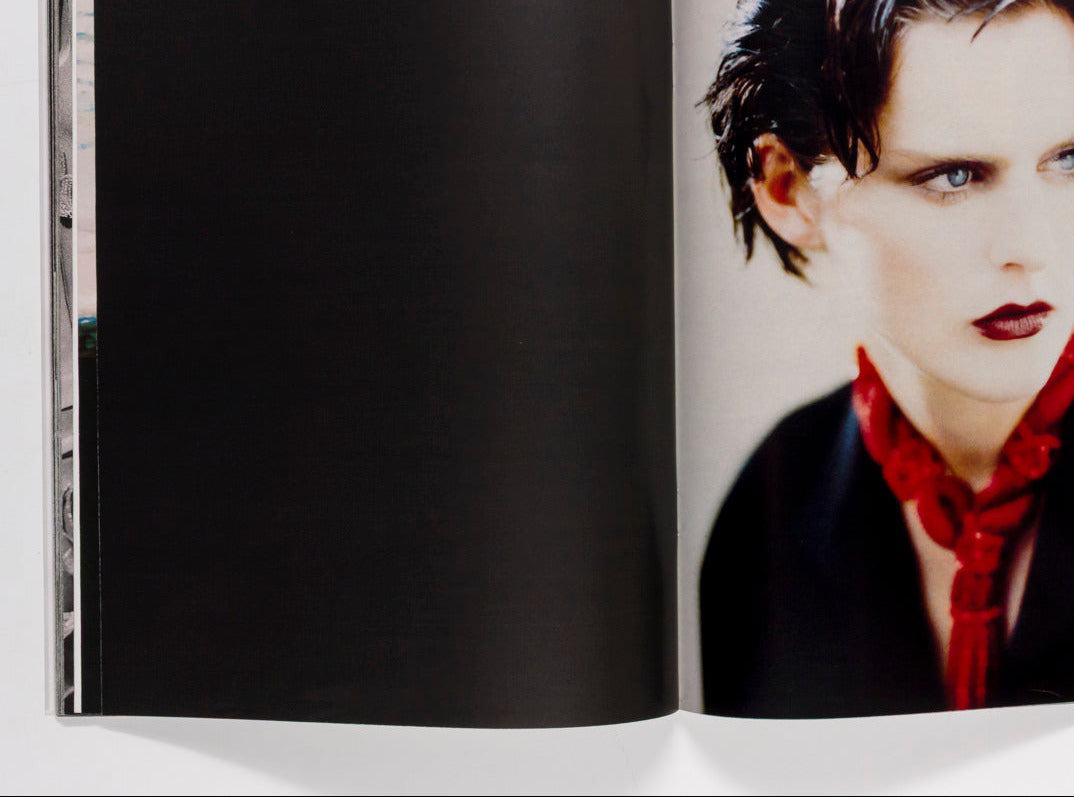 An open book or magazine spread, with the left page in black and the right page showing a portrait photograph of a woman with short dark hair, blue eyes, red lipstick, and a red neck scarf against a light gray background.